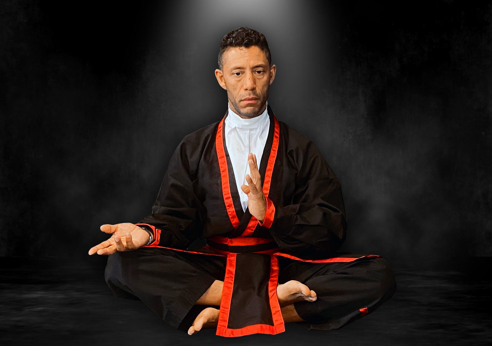 PKJN Nathan, dressed in a black martial arts uniform with red trim, sits cross-legged in a meditative pose with one hand open and the other held vertically in a focused gesture, against a dark smoky background illuminated by a soft spotlight.