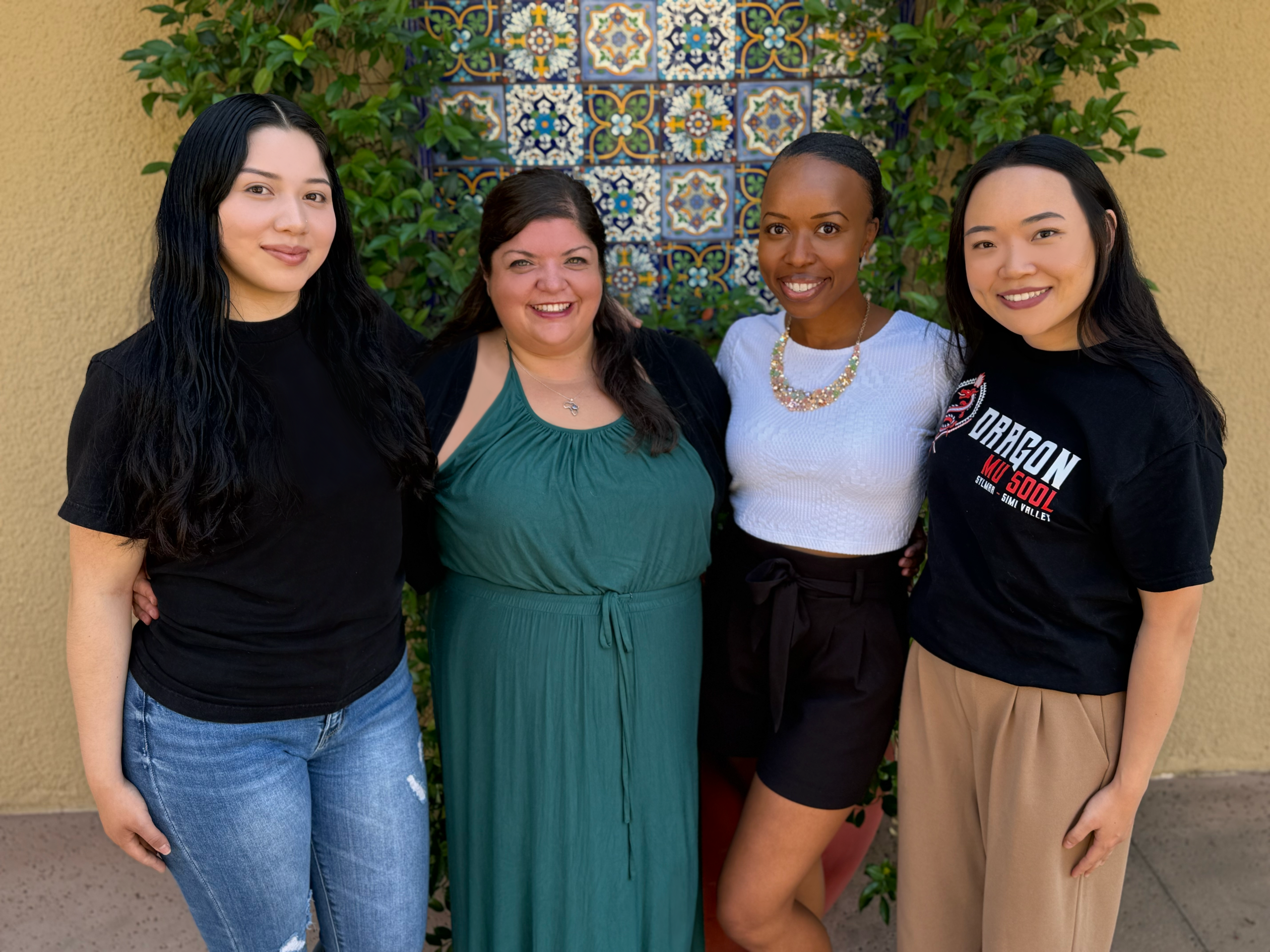 Dragon Mu Sool Client Services Photo The Dragon Mu Sool Client Services team poses together outdoors in front of a colorful tiled wall and greenery. The group of four women stands side by side, smiling warmly. They wear a mix of casual and semi-formal outfits, including one team member wearing a black Dragon Mu Sool T-shirt.