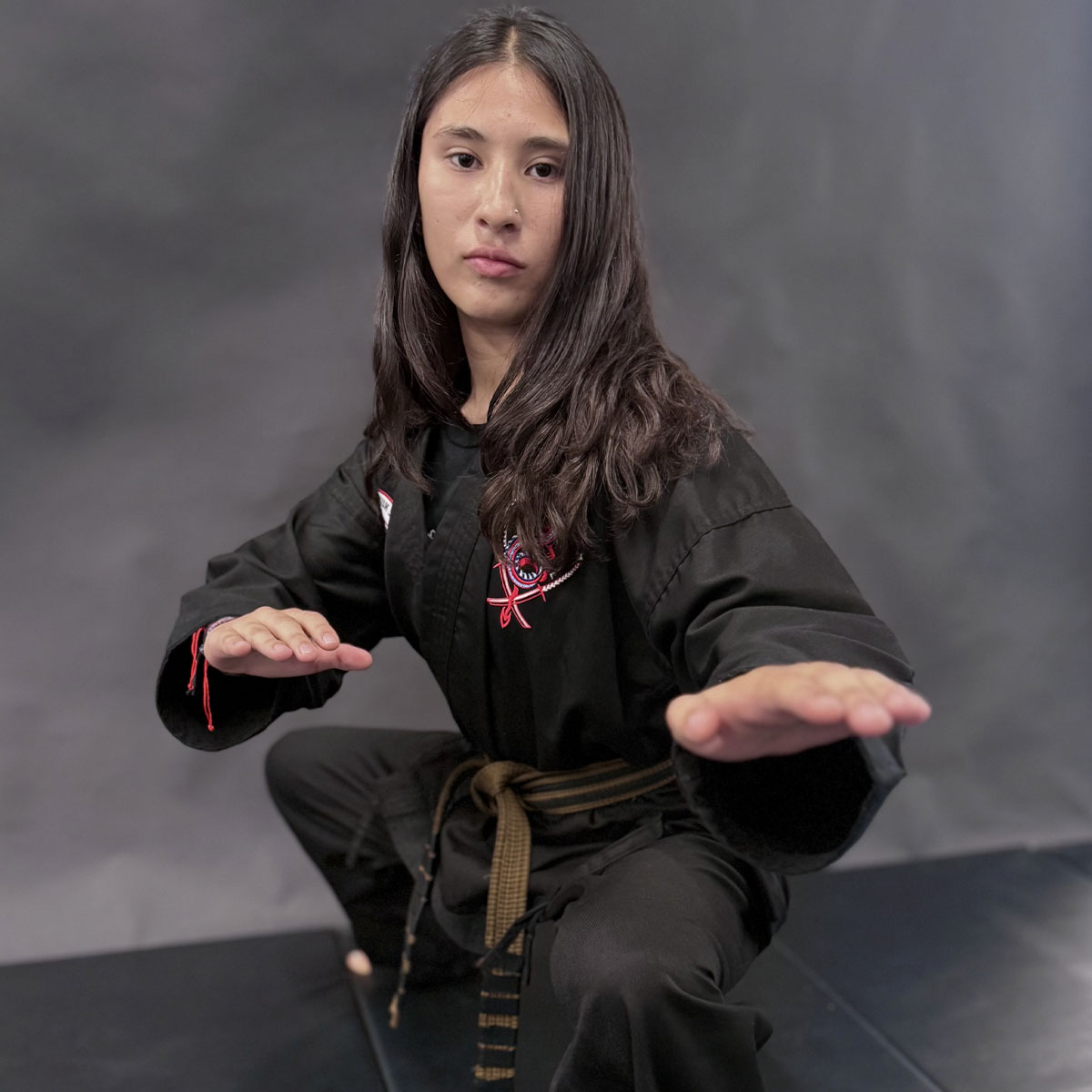 JKN Adriana, A young martial artist in a black uniform with a brown belt poses in a low stance, hands extended in a defensive position. The uniform features the Dragon Mu Sool logo embroidered on the chest. The background is a neutral gray studio backdrop.
