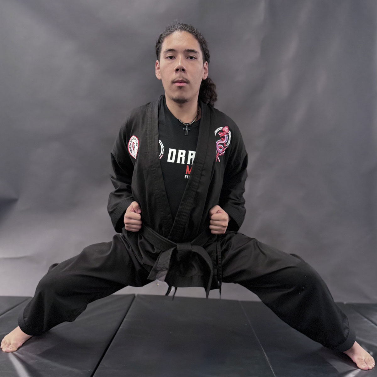 JKN Josiah, A martial artist performs a wide horse stance on padded flooring while wearing a black uniform with a black belt. The uniform and shirt feature the Dragon Mu Sool logo and text in red and white. The person looks forward with focus against a gray studio backdrop.