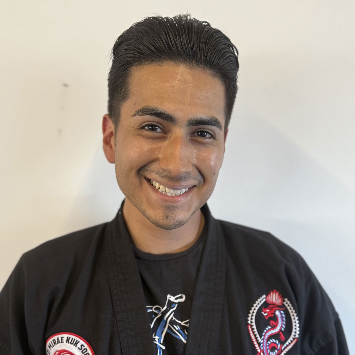 JKN Olyn, A smiling martial artist wearing a black uniform with Dragon Mu Sool patches on the chest and shoulder. The person stands against a plain light background, exuding confidence and warmth.