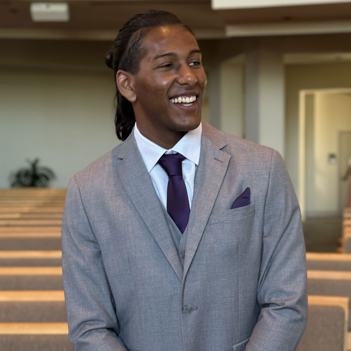KSN Cyrus, dressed in a light gray suit with a deep purple tie and pocket square, smiles warmly while standing indoors. The background shows rows of seating and soft lighting, giving the scene a formal yet relaxed atmosphere.