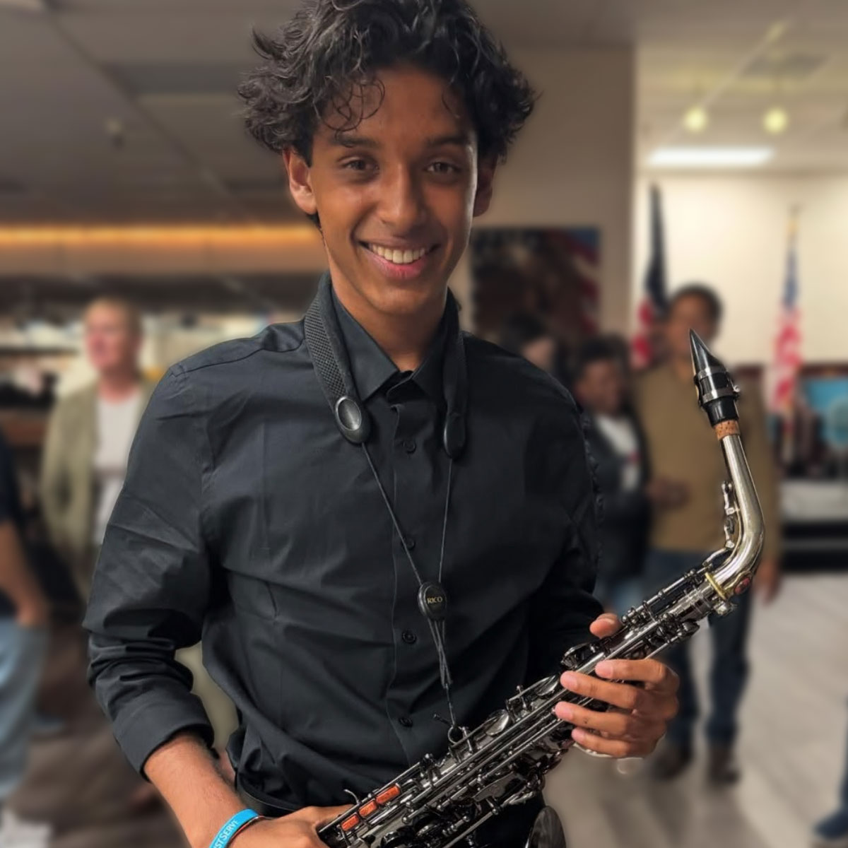 KSN Hunter smiles while holding a saxophone in a warmly lit indoor setting. He’s dressed in a black button-up shirt, with a saxophone strap around his neck. The background shows several people and American flags slightly out of focus, suggesting a formal or celebratory event.